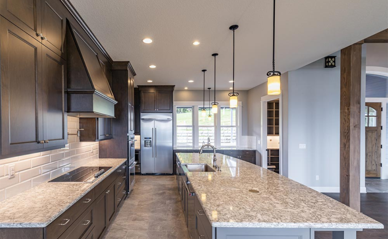 A modern kitchen with a large quartz countertop island, dark wood cabinets, stainless steel appliances, pendant lights, and natural light from large windows.