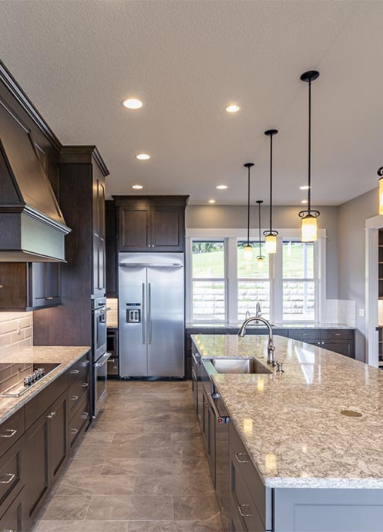 A modern kitchen with a large quartz countertop island, dark wood cabinets, stainless steel appliances, pendant lights, and natural light from large windows.